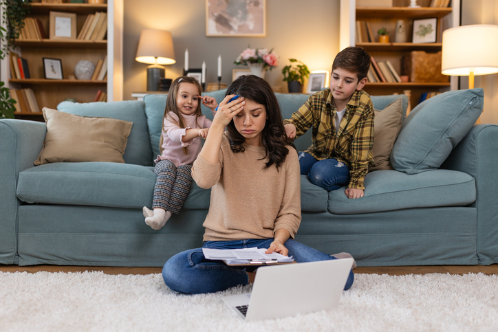 A stressed mother using a laptop while her daughter and son prod her from the couch behind her.