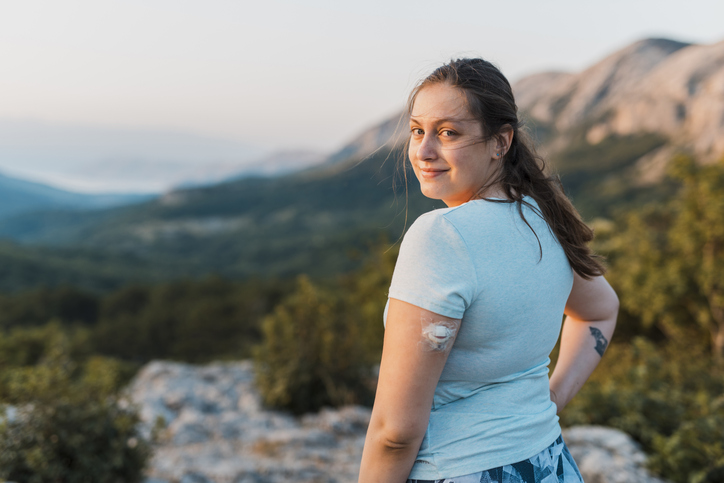 A woman wearing a glucose monitor stares confidently at the camera while on top of a mountain.