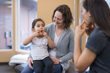 A little girl sits on a woman's lap while mimicking the face made by a speech pathologist seated across from her.