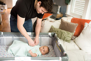 A man places a baby, wearing a sleep sack, in a bassinet. 