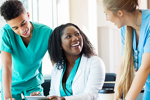 Two young, white adults in scrubs stand on either side of a seated African American women wearing a white lab coat. All three are talking and laughing.