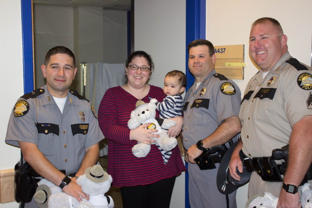 Three Kentucky State Troopers deliver a teddy bear to a young patient and mom.