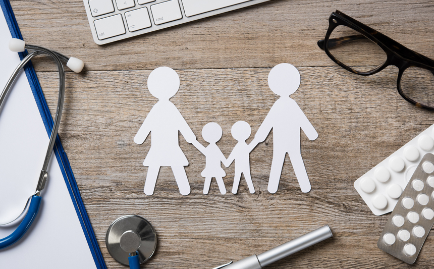 A set of paperdolls representing two parents and two children near a computer keyboard, stethoscope and pill blister packs.