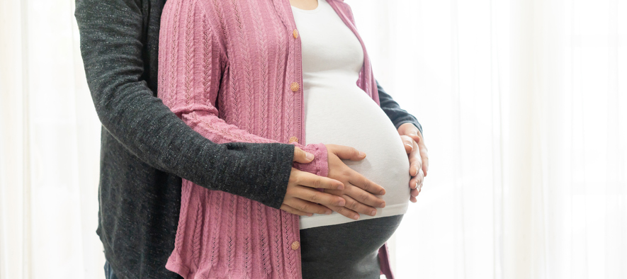 A pregnant woman is hugged by her spouse from behind.