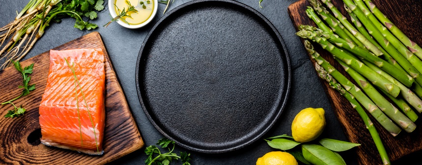 A raw piece of salmon lies on a cutting board, surrounded by asparagus and herbs.