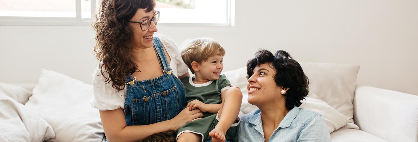 Two smiling women and a toddler boy sit together on a white sofa