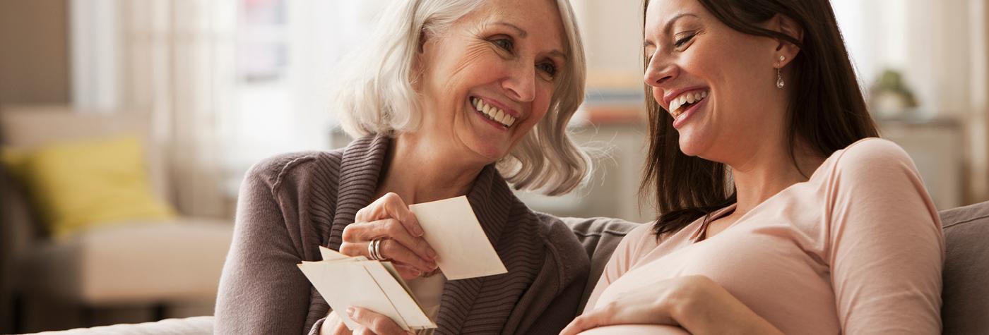 A young, pregnant woman sits next to a smiling older woman and laughs as they look at photographs together.