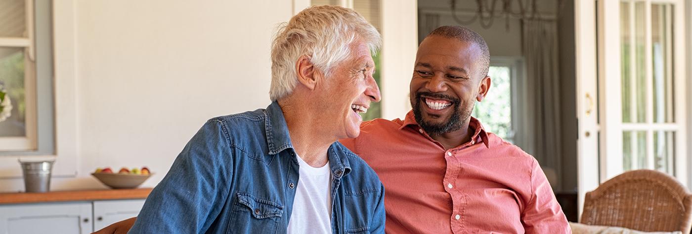 Two middle-aged men laugh with one another while sitting side by side on a couch.