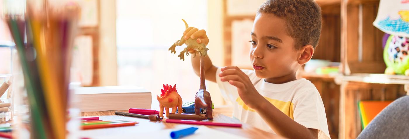 A young African-American boy plays with toy dinosaur figurines in a well-lit playroom. 