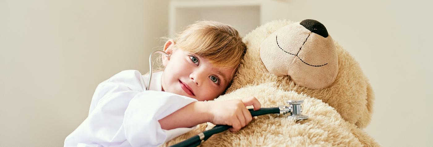 A little girl dressed in a white coat uses a stethoscope on a large teddy bear. 