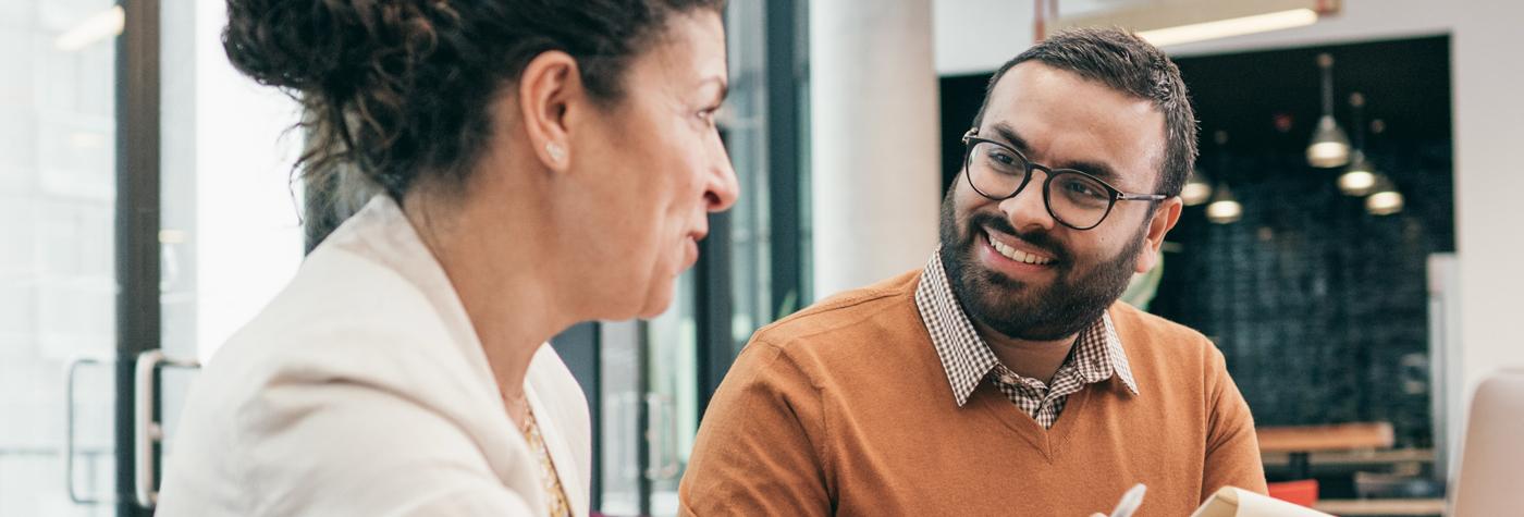 A man and woman dressed in business casual attire smile at one another while having a discussion. 