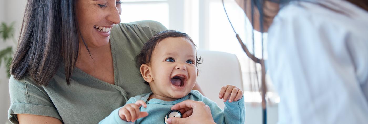 A baby, wearing a light blue, long-sleeved onesie, sits on its mothers lap and laughs while a female doctor wearing a white coat uses her stethoscope to listen to the baby's heart.