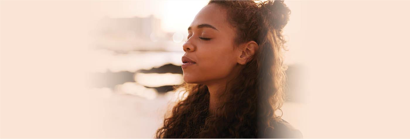 A woman with brown skin and long, wavy brown hair meditates while sitting on rocks along a shoreline.