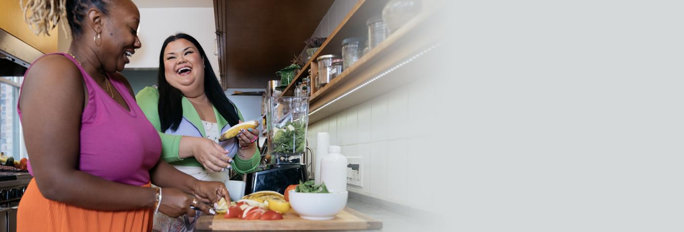 Two women talk and laugh together while standing at a kitchen counter preparing vegetables.