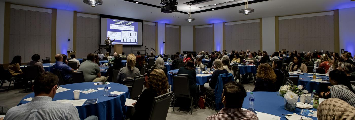 A researcher stands on a stage and makes a presentation to a room full of audience members.