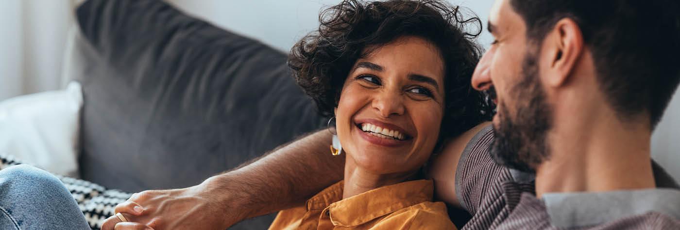 A man and woman sit together on a couch and smile at one another. 