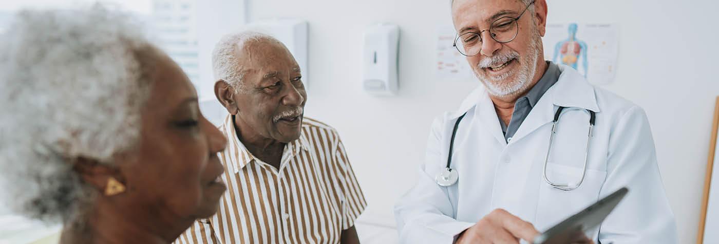 An older Black man and woman meet with a doctor -- also an older man with a gray goatee -- who is wearing a white coat, has a stethoscope draped around his neck, and is showing the couple something on a tablet computer.