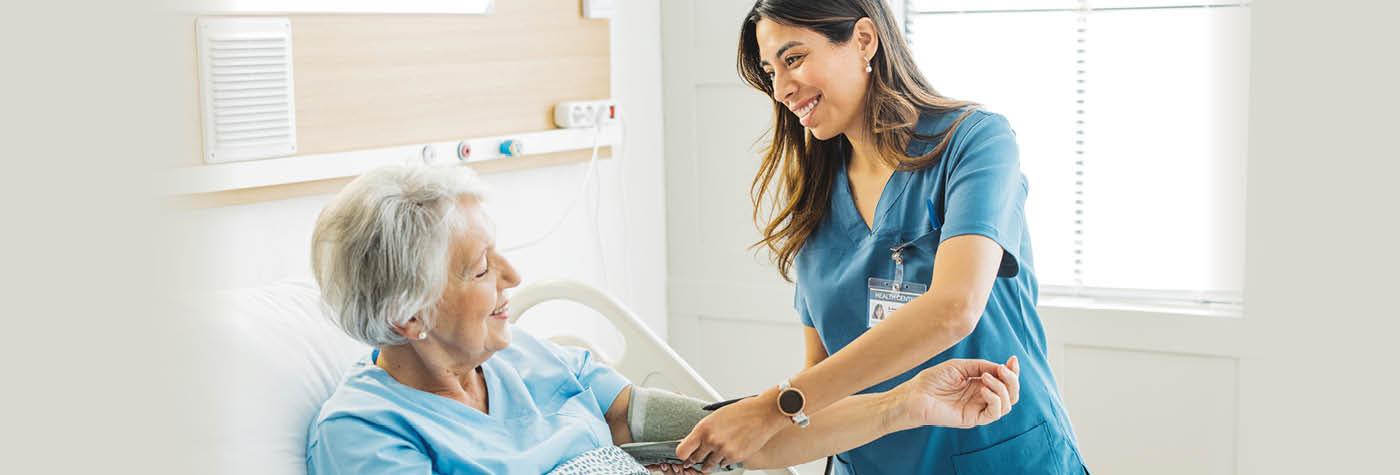 An older woman sits up in a hospital bed while a smiling female in blue scrubs uses a blood pressure cuff on her arm.