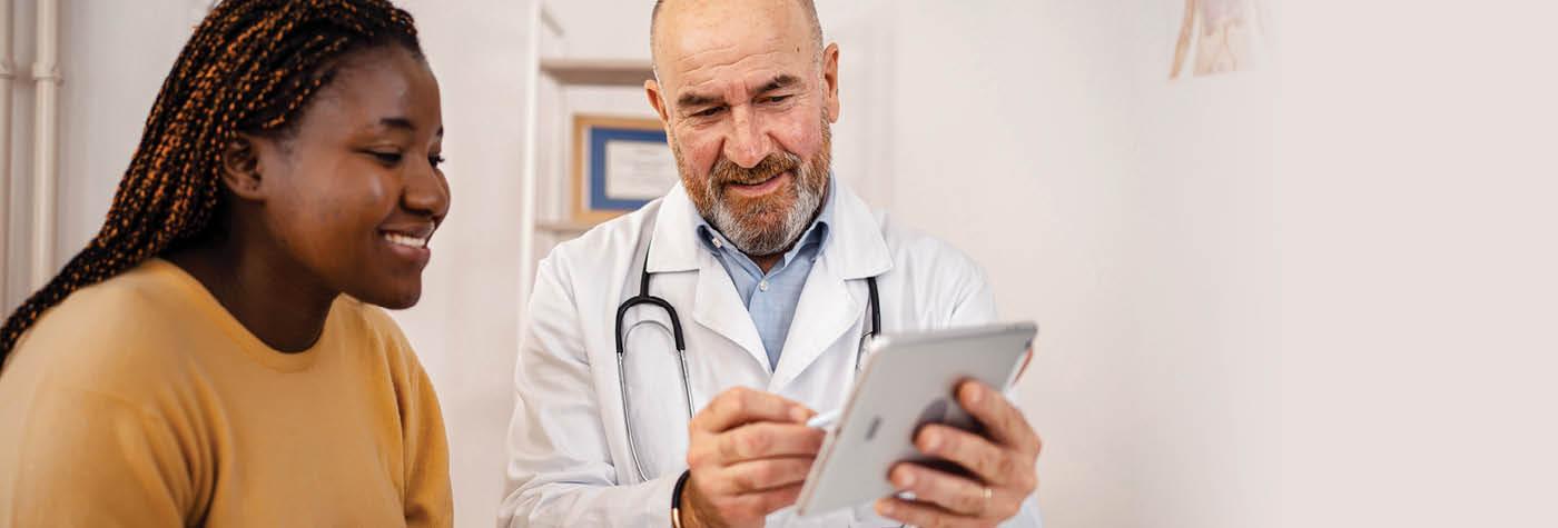 A male physician wearing a white coat discusses something on a tablet computer with his patient, a young black woman wearing a yellow sweater.