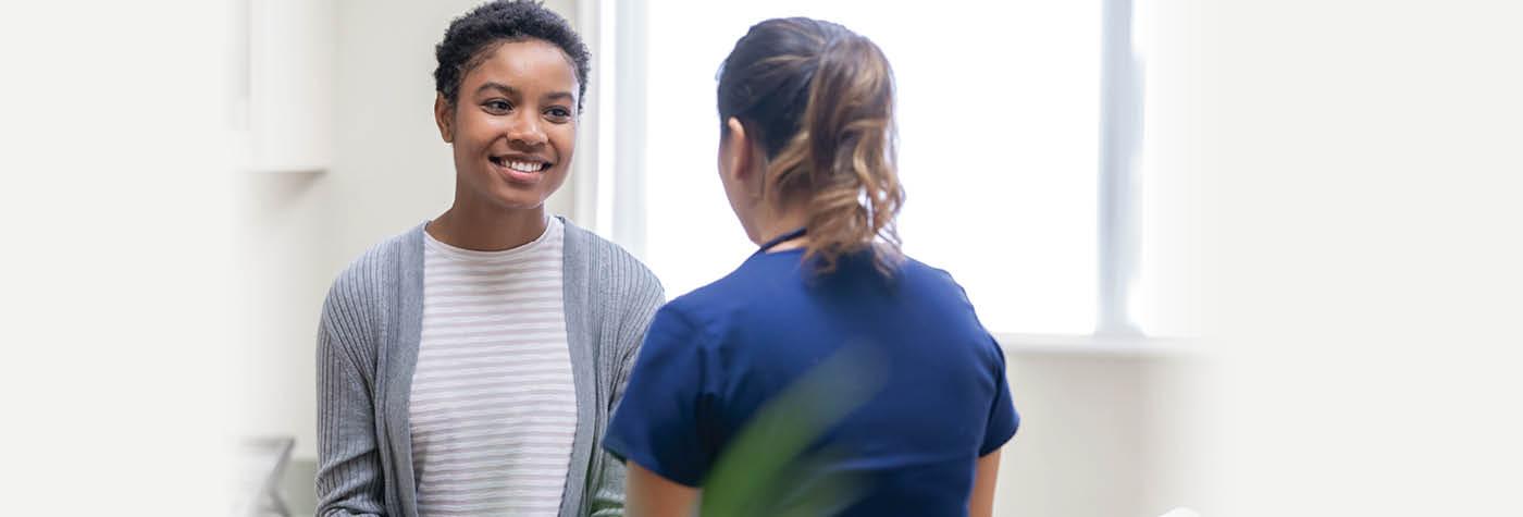 A female patient sits across from a female provider wearing blue scrubs. She is smiling and speaking to the provider.