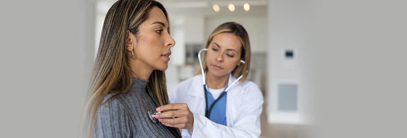 A woman wearing a gray sweater sits in an exam room as a female doctor wearing a white coat and light blue scrubs uses a stethoscope to listen to her chest.