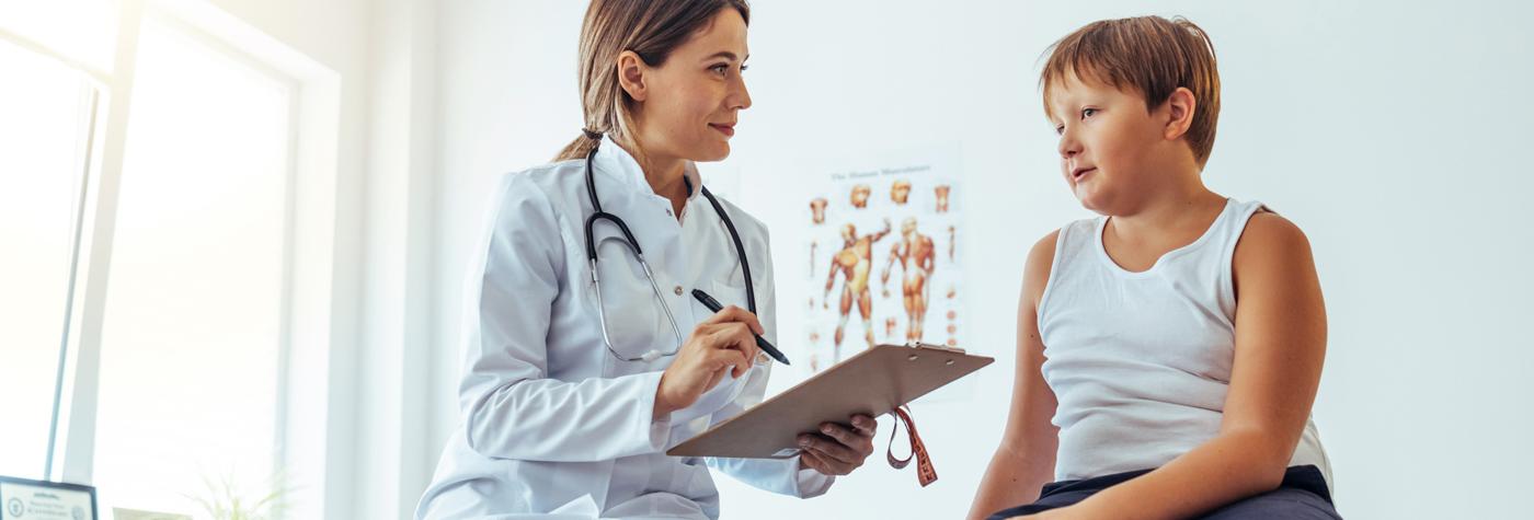 A female provider wearing a white coat with a stethoscope draped around her neck holds a clipboard and pen while talking to her patient, a preteen boy wearing a white undershirt.