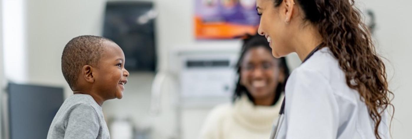 A young African-American child smiles as he talks to his doctor, a woman who is also smiling. His mother sits in the background, smiling as she looks on. 