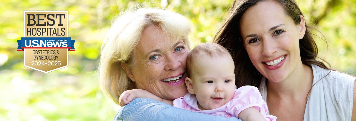 An older woman and younger woman stand close together while holding a smiling baby wearing a pink blouse. A badge superimposed over the image reads US News and World Report Best Hospitals, Obstetrics & Gynecology, 2024-2025