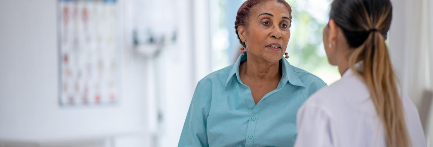 An older woman wearing a light blue blouse speaks with her doctor, a woman with a long ponytail and wearing her white coat.