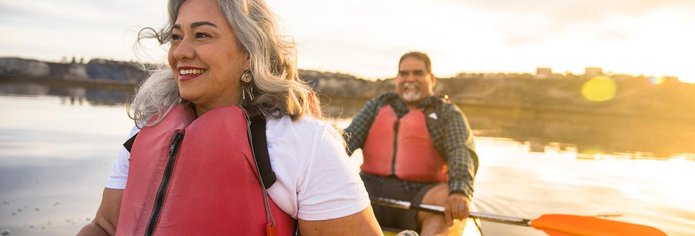 A man and woman, both wearing life vests, smile as they float on a lake in a two-person canoe.