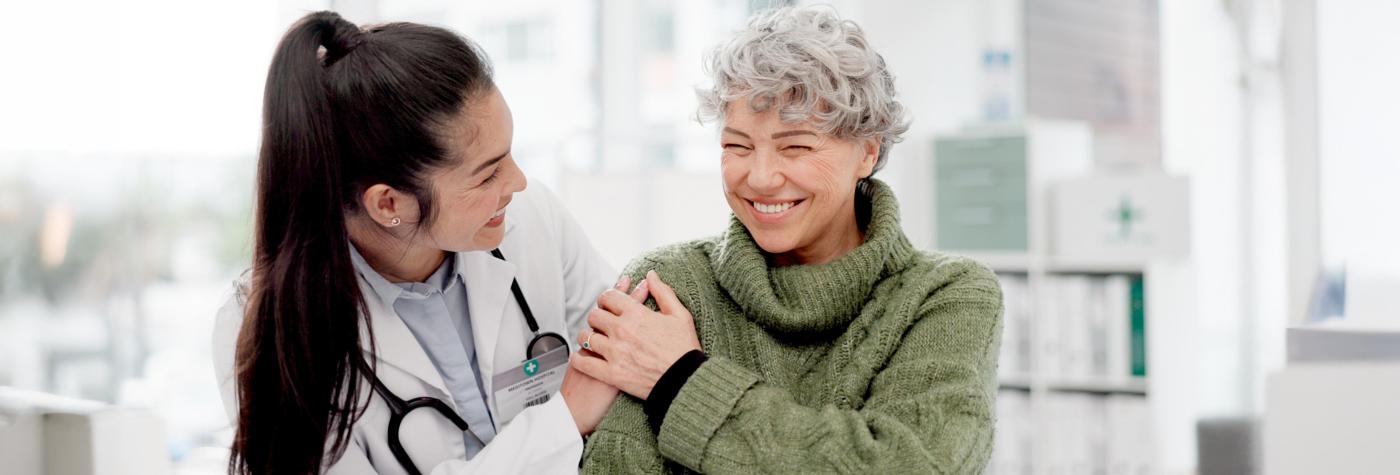 A female healthcare worker with long, dark hair in a ponytail and wearing a white coat with a stethoscope around her neck smiles as her hand is patted by the hand of a female patient with a short, grey haircut wearing a green turtleneck sweater.