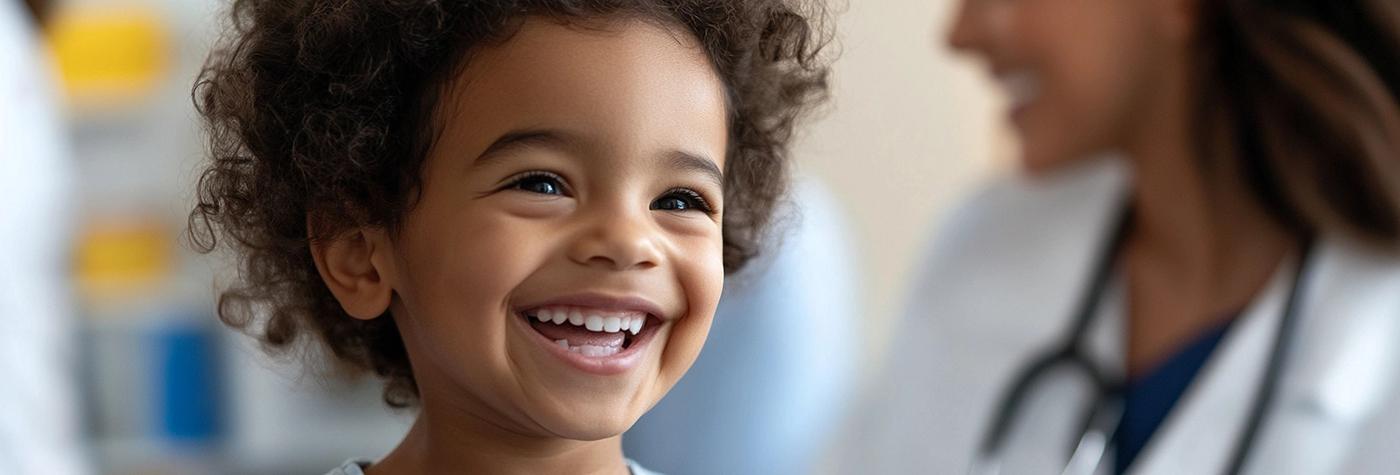 A young child smiles while his provider, a woman wearing a white coat and stethoscope draped around her neck, smiles out of focus in the background.