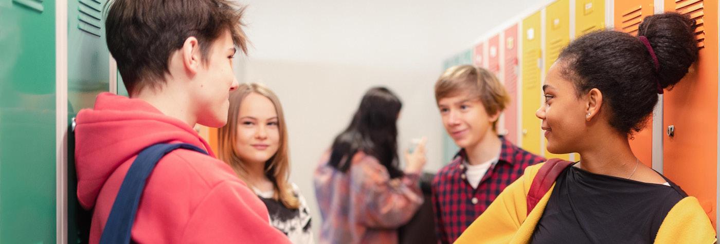 A group of teens gather next to a bank of lockers in a school hallway.