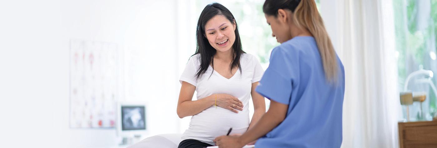 A pregnant woman wearing a white tee shirt sits on an exam table and speaks with a woman wearing light blue scrubs. 