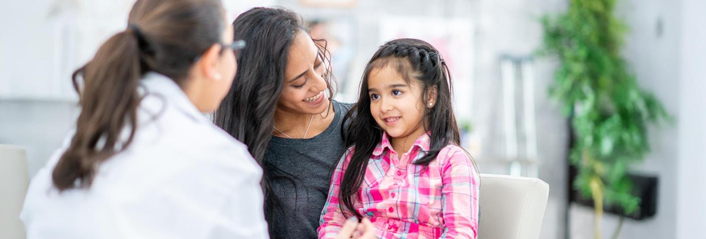 A female doctor, wearing a white coat, talks to a young girl dressed in a pink plaid shirt, who is seated on the lap of her mother, a woman with tan skin and long black hair wearing a dark grey shirt. 