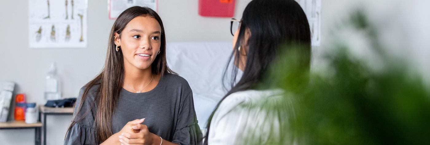 A teenage girl with long, dark brown hair and tan skin, wearing a dark grey shirt, talks to her doctor, a woman with long black hair who we see over the shoulder.