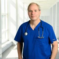 Dr. Michael Anstead, a middle-aged white man with grey hair, stands in the hallway of UK HealthCare wearing blue scrubs with a stethoscope around his neck.