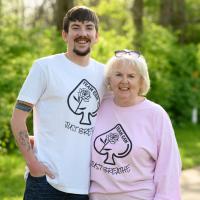 Cameron and his grandmother, an older white woman with white hair, stand and smile side by side. She is wearing a pink long sleeved ‘Team Cam’ shirt with her spectacles on her head.