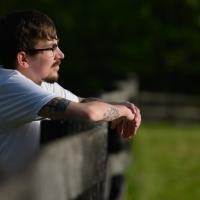 Cameron stands turned to his side, with his hands on the border fence of the park as he stares into the horizon.