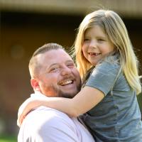 Sarah Beth is held up and embraced by her father, Scott Ferguson, a middle-aged white man with short brown hair wearing a light pink shirt.