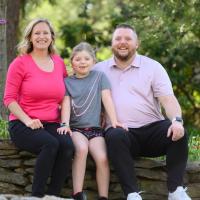 The Ferguson family sits outside and smile together. Sarah Beth is seated between her father, Scott and her mother, Erin, a middle-aged white woman with blonde hair, who is wearing a pink shirt.