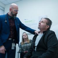 Dr. Andrew Kolodziej, left and standing, holds the shoulder of heart-transplant patient Ron Kelly, who's seated. Between them, viewable under Dr. Kolodziej's arm, is Ron's partner Deborah, also seated.