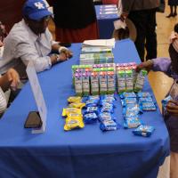 A little girl wearing a purple sweater takes a juice box from a table with a blue table cloth. Sitting across from her at the table are a man and a woman.