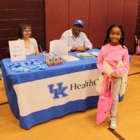 A young girl wearing a pink sweater stands in front of a table with a blue and white, UK HealthCare-branded tablecloth. Two adults -- one man and one woman -- sit at the table.