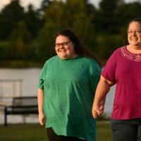 A candid photo of Abby and her mother, Jamie, smiling as they walk hand in hand in front of the water. Jamie is an older white woman with long brown hair. She is wearing a magenta top that has an embroidered pattern around the collar, and a pair of glasses.