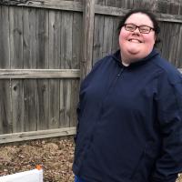 A photo of Abby smiling in front of a wooden fence while out on a walk.