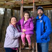 Addison stands outside with his family, including his wife, a middle-aged white woman with brown hair wearing a grey jacket and a pink flannel shirt, and their daughter, a young white girl with a purple jacket and pink pants.