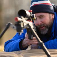 Addison lays on the ground in the shooting position, with his hand on the trigger as he looks through the rifle scope.