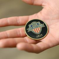 Dr. Kluemper holds out his hand to reveal the medallion given to him by Addison. The medallion is gold with a black background with the US Rifle team emblem and the year 2021 displayed in the front.