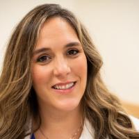 Dr. Maureen O’Shaughnessy, a young-looking white woman with long, wavy brown hair, smiles at the camera. She is sitting and wearing a white lab coat. We see her from the shoulders up.